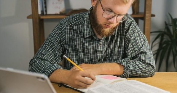 A bearded man with glasses looks at the easiest Bible to read and understand and makes notes with a pencil