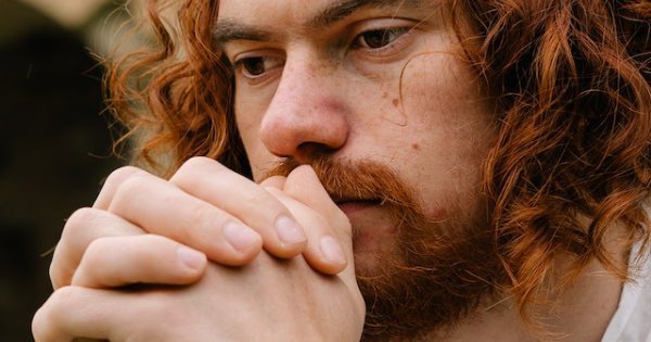A man with red wavy hair and a beard clasps his hands together in prayer. He looks confused because he does not know how to pray to God.